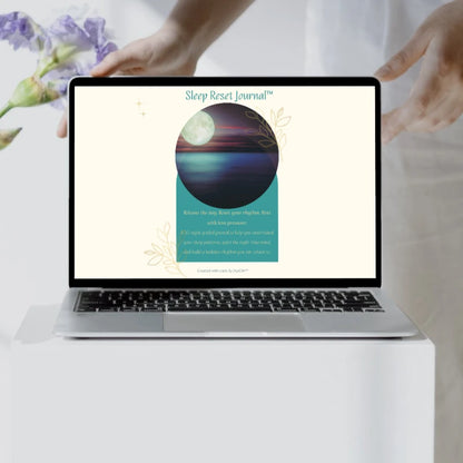 Sleep Reset Journal™ cover displayed on a laptop on a white table beside purple flowers.